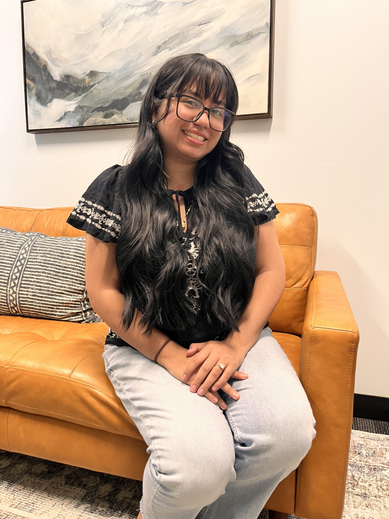 A woman with long wavy black hair and glasses smiles while sitting on a tan leather couch. She is wearing a black embroidered blouse and light blue jeans, with her hands gently folded in her lap. A large abstract painting hangs on the wall behind her.
