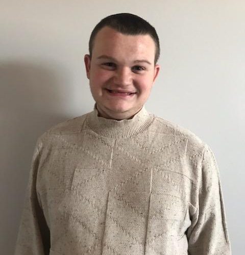 A young man with short dark hair smiles while standing in front of a plain light-colored wall. He is wearing a beige textured sweater with a geometric pattern.