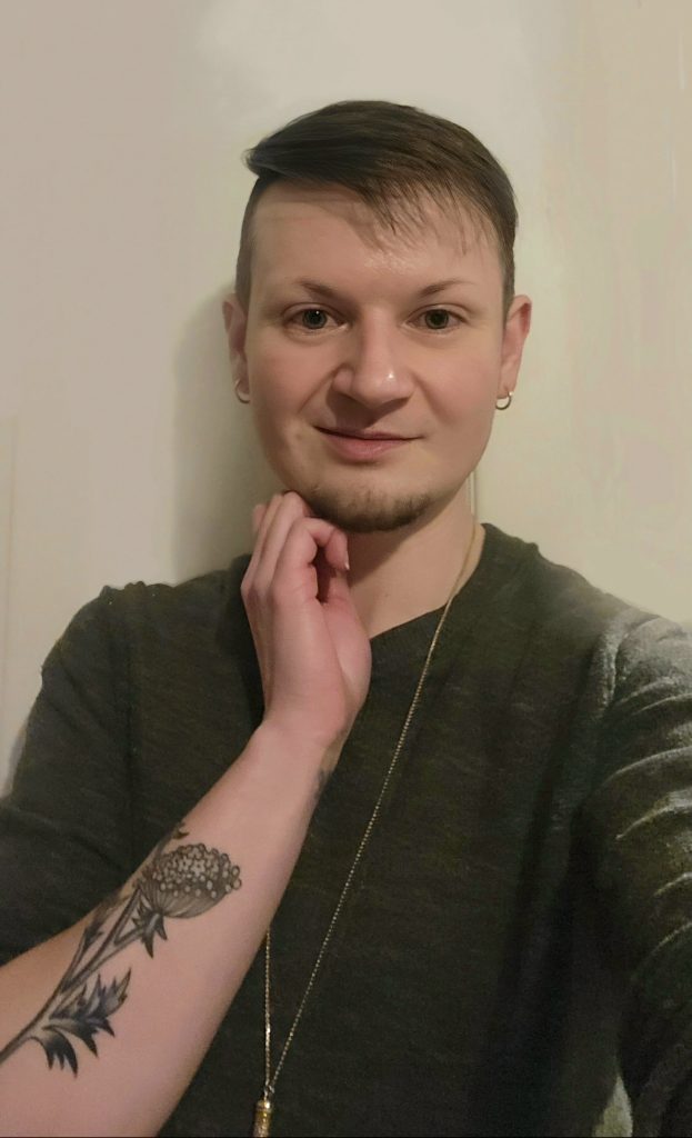 A person with short, styled brown hair and light skin poses indoors against a light-colored wall. They are wearing a dark green top, a long necklace, and small hoop earrings. One hand gently touches their chin, revealing a detailed black ink floral tattoo on their forearm. They are smiling slightly and looking directly at the camera.